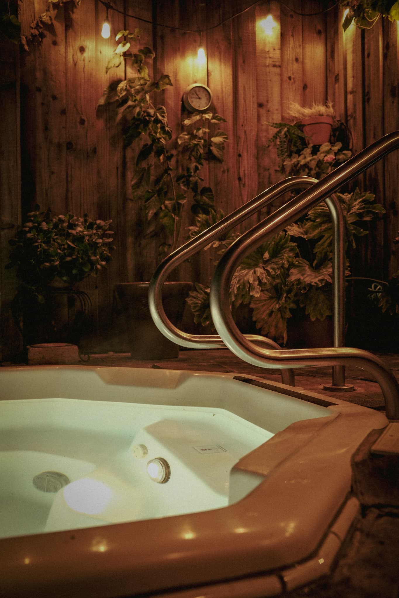 A private outdoor hot tub glowing beneath warm orange lights, cedar, and plants at night