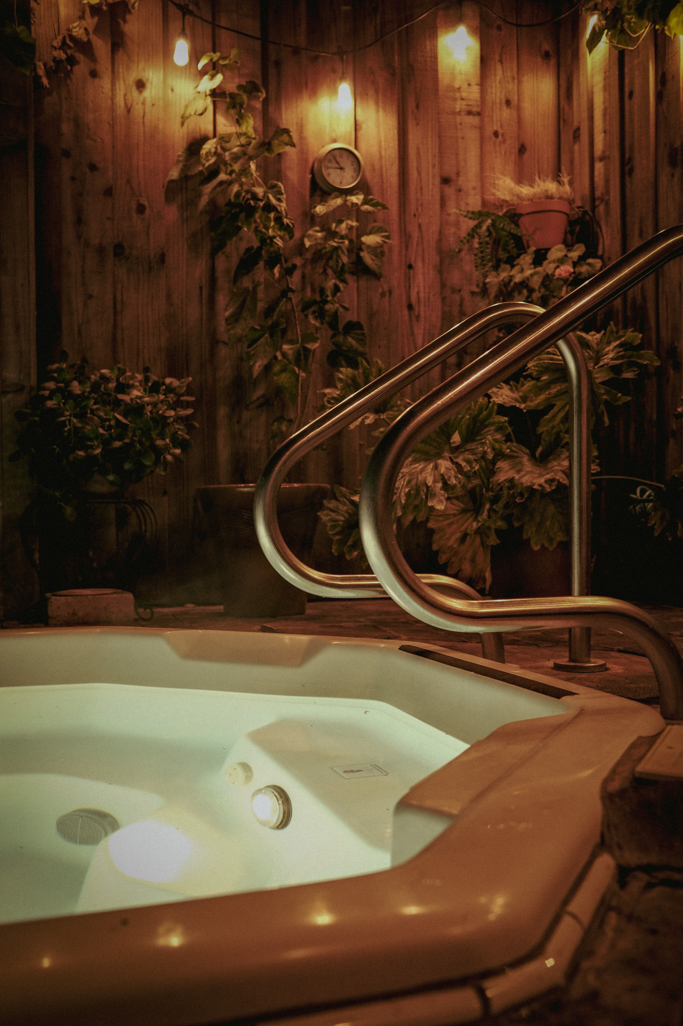 A private outdoor hot tub glowing beneath warm orange lights, cedar, and plants at night
